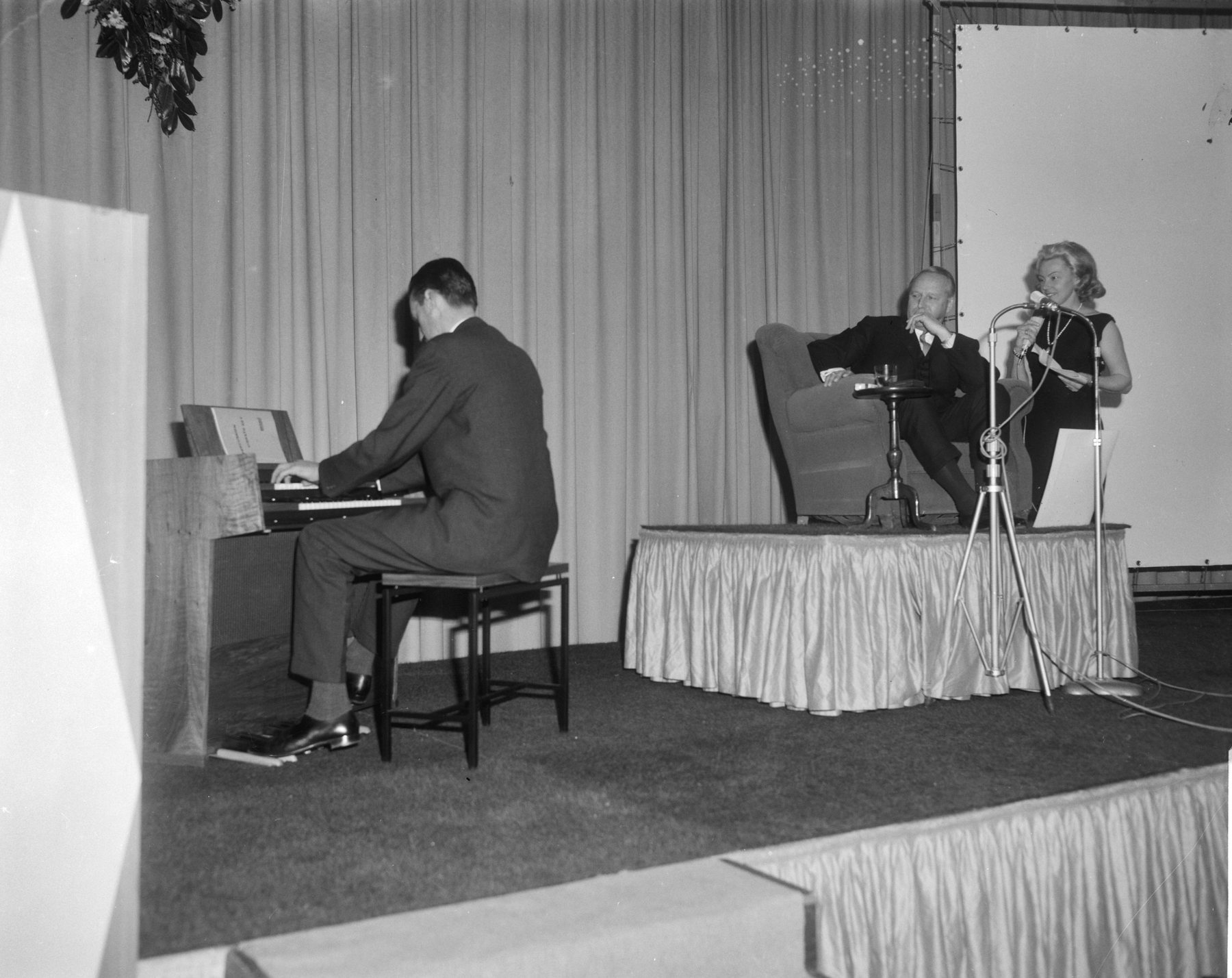Een pianist speelt op een podium, een man zit in een fauteuil en een vrouw staat bij de microfoon