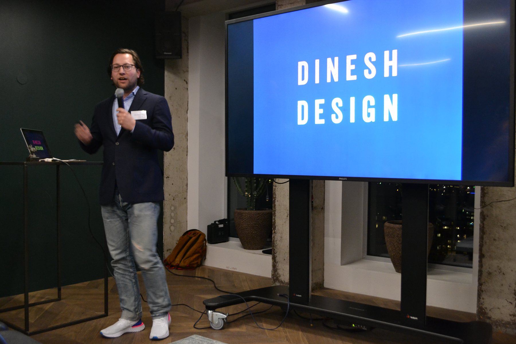 Een man spreekt met een microfoon naast een scherm met de tekst Dinesh Design