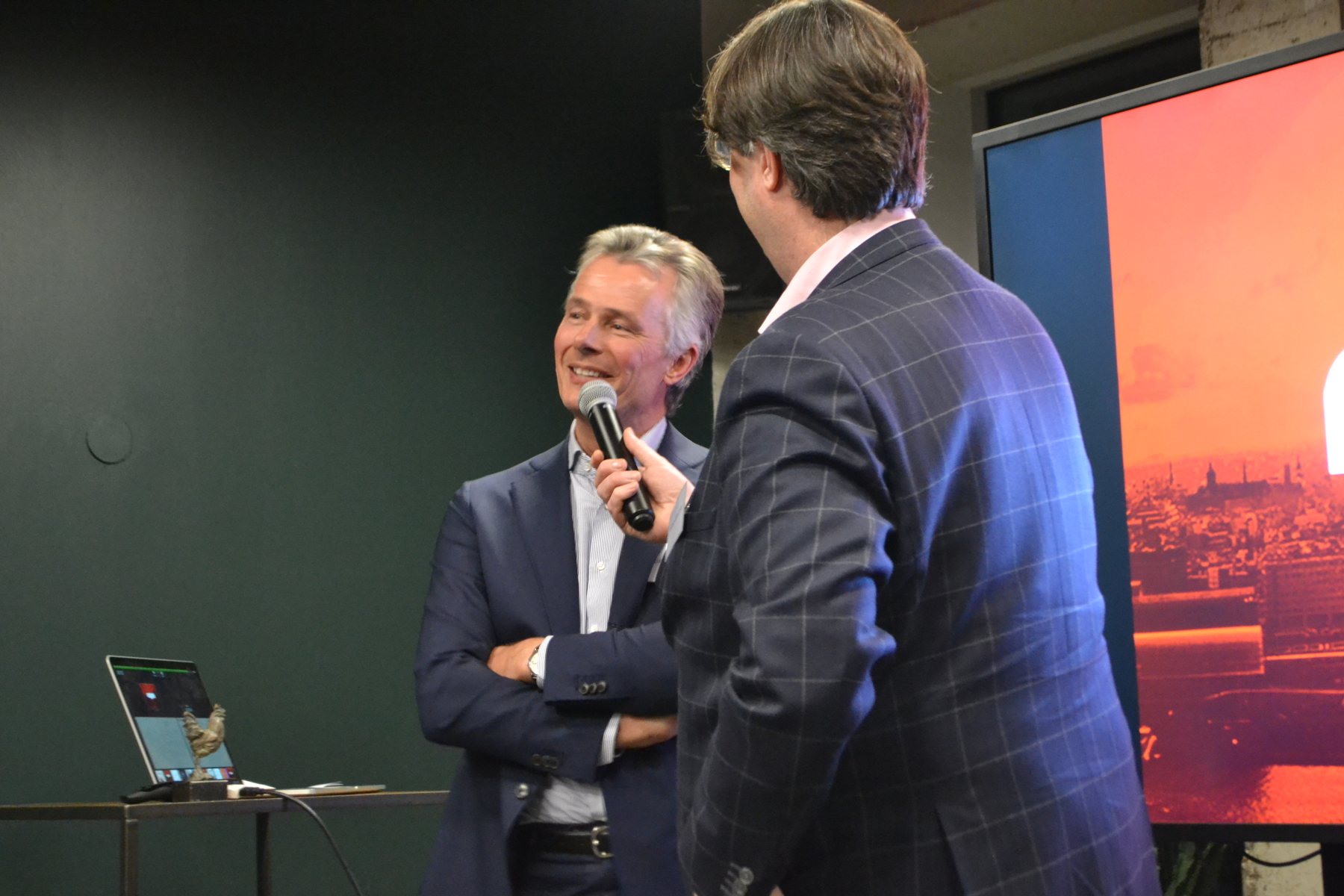 Twee heren in pak in gesprek met een microfoon, voor een groot scherm